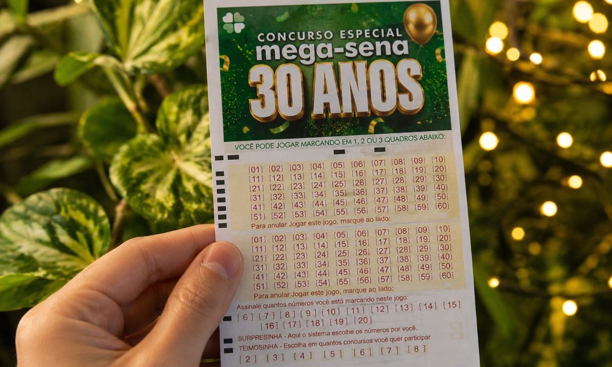 Mão segurando volante da Mega-Sena 30 anos com fundo de planta atrás