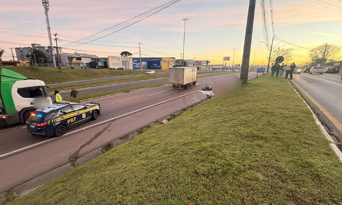 Foto mostra, de longe, movimentação da PRF no local do acidente que matou motociclista que bateu de frente com caminhão na Rodovia do Xisto. A pista sentido Curitiba está totalmente interditada. O dia está no início do amanhecer. O corpo do motociclista aparece caído no asfalto, ao fundo, ao lado do caminhão