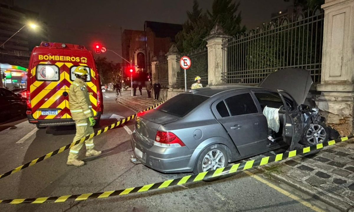 Um motorista optou por jogar o próprio carro contra um muro no Batel. Imagem mostra uma ambulância do Corpo de Bombeiros na Avenida do Batel. O carro está parado na calçada com a parte da frente destruída. O carro é cinza.