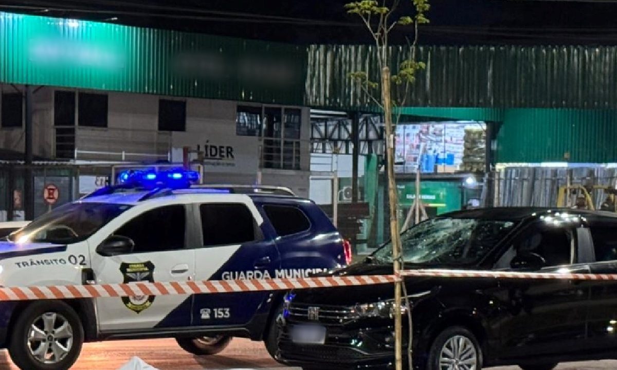 Foto tirada durante à noite mostra corpo de homem que morreu atropelado na Linha Verde caído na frente do carro envolvido no acidente. Corpo está coberto por pano branco e foi borrado em edição. Viatura da Guarda Municipal aparece parada ao lado do carro preto do acidente e do corpo do homem. Movimento de carros no local é intenso.