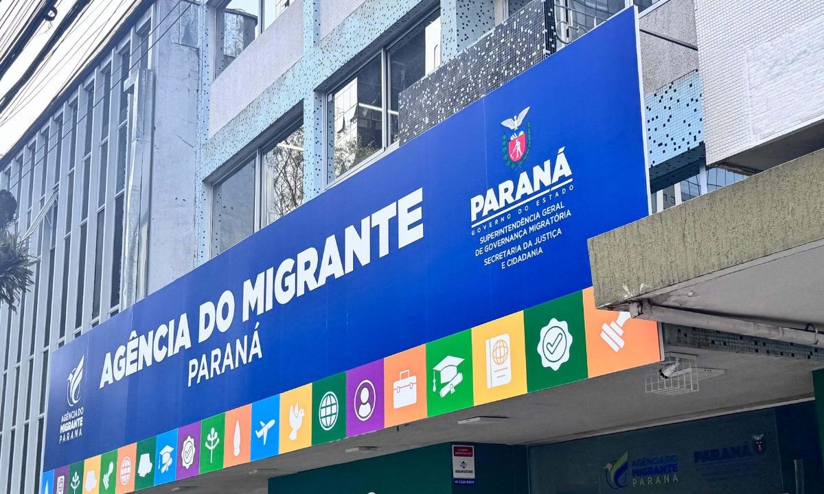 Fachada da Agência do Migrante Paraná, em Curitiba, com placa azul do Governo do Estado na entrada do prédio.