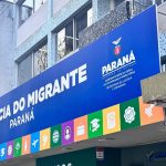 Fachada da Agência do Migrante Paraná, em Curitiba, com placa azul do Governo do Estado na entrada do prédio.