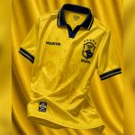 Imagem mostra a camisa do Paraná Clube 2026 em fundo amarelo, inspirada no uniforme da Seleção Brasileira campeã do mundo em 1994. A peça tem predominância da cor amarela, detalhes em azul na gola e mangas, escudo estilizado do clube e referências visuais ao tetra mundial