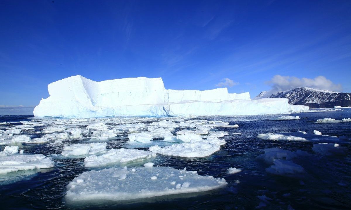 Grande iceberg cercado por placas de gelo flutuando no oceano pelo derretimento na Antártida sob céu azul intenso.