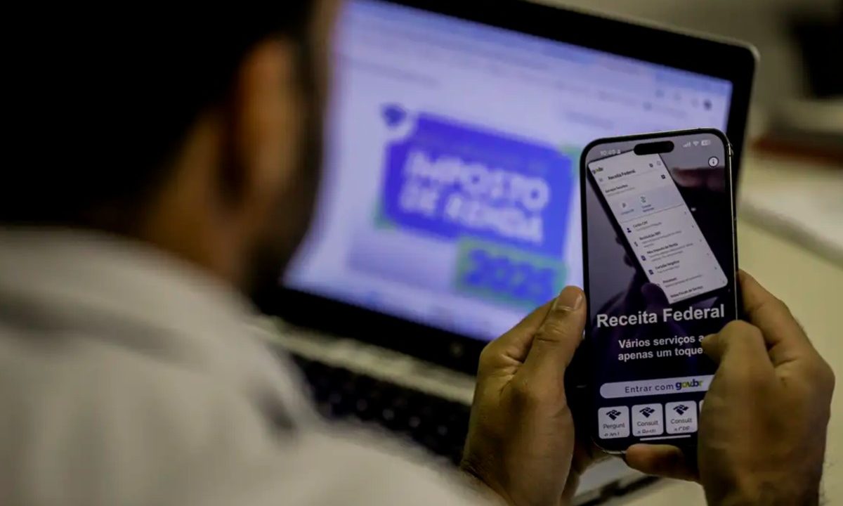 Homem segura um celular com o aplicativo da Receita Federal aberto na tela. Ao fundo, desfocado, há o escrito "Imposto de Renda", em alusão à abertura da consulta do primeiro lote de restituição