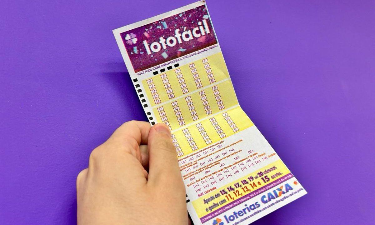 Mão segunrando o volante da Lotofácil 3685 com um fundo roxo.