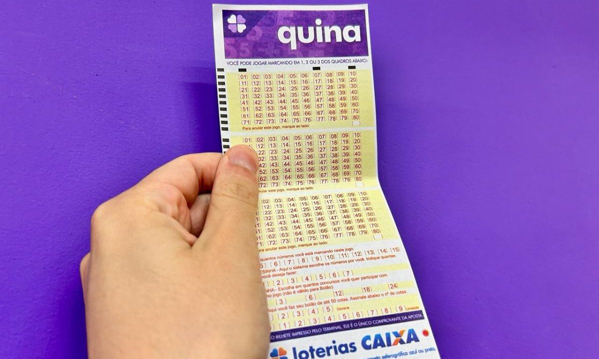 Mão segurando o volante da Quina 7027 em frente a um fundo roxo.