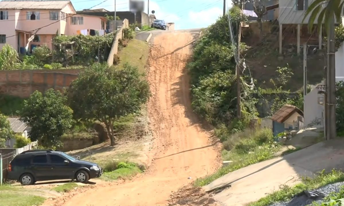 Vista da Rua Nossa Senhora das Graças, em Almirante Tamandaré. A imagem mostra uma subida extremamente íngreme em solo de terra avermelhada e saibro, cercada por casas simples e vegetação. Um carro preto está estacionado na base da ladeira, evidenciando o declive acentuado.