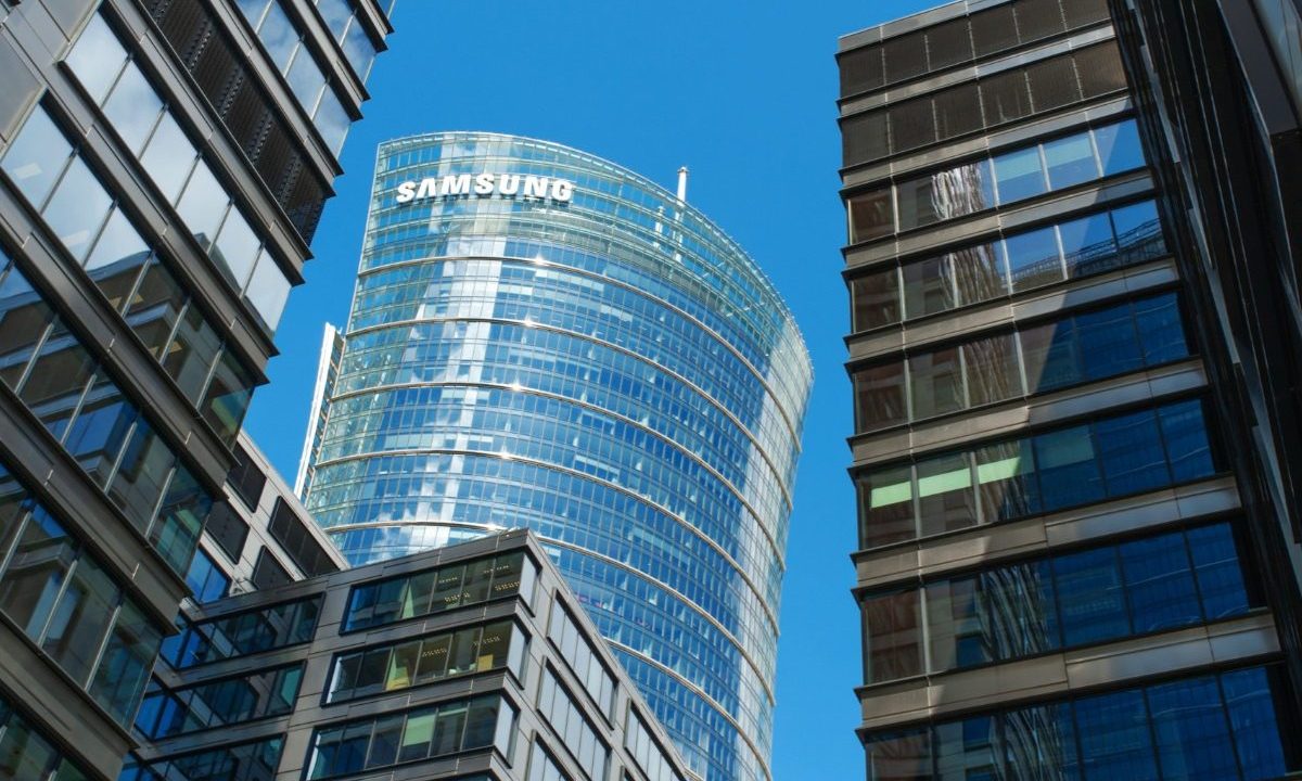 Na imagem aparece o prédio da Samsung, toda em vidro, em formato circular e um céu azul ao fundo, além de prédios ao redor.