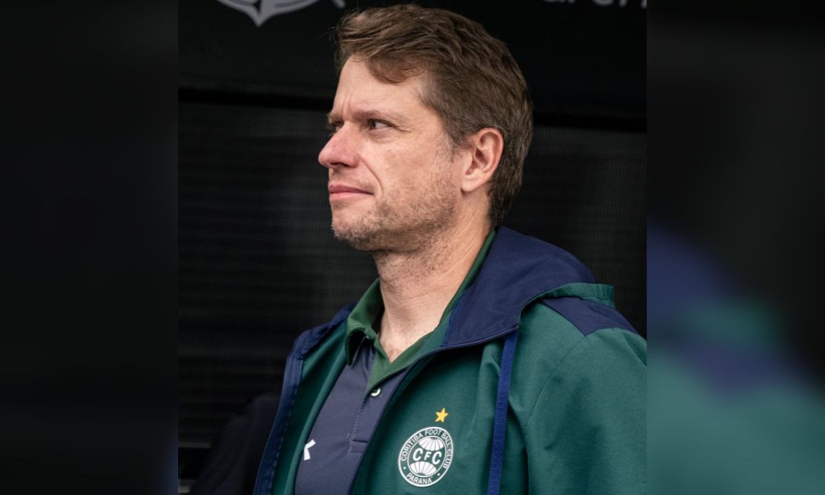 Na imagem, Fernando Seabra aparece de perfil, olhando para o lado, com expressão séria. O treinador veste um agasalho verde e azul do Coritiba, com o escudo do clube no peito, em ambiente ligado ao estádio.