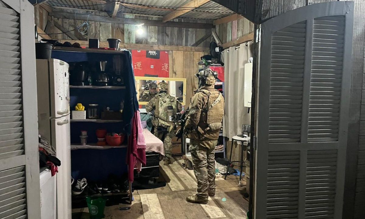 Na imagem aparecem dois agentes da polícia utilizando fardas de combate camufladas, em uma casa de madeira. A porta estilo colonial aberta, uma geladeira ao canto, uma estante de madeira e diversos objetos espalhados. serie-crimes-litoral-assassinatos-matinhos-vitimas 3