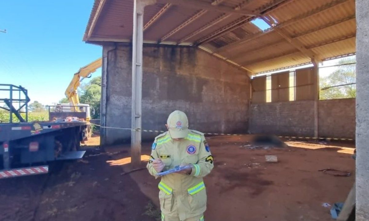 Foto mostra bombeiro fardado anotando informações em uma prancheta em primeiro plano. Ao fundo, no chão, está o corpo do trabalhador que morreu coberto por uma lona preta. No alto, é possível ver o telhado do barracão quebrado.