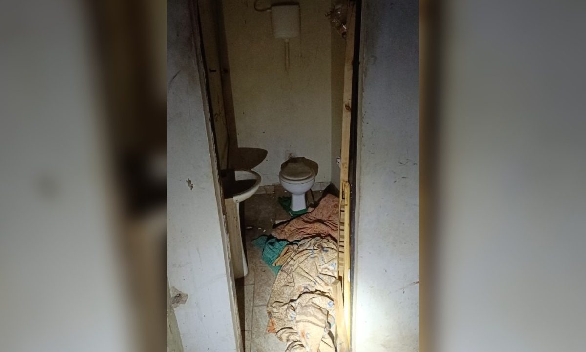 Porta arrombada e aberta, para um banheiro pequeno e sujo, apenas com privada e pia, com cobertas no piso.