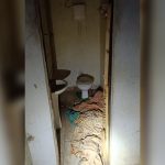 Porta arrombada e aberta, para um banheiro pequeno e sujo, apenas com privada e pia, com cobertas no piso.