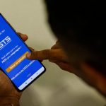 Foto em plano detalhe, tirada por cima do ombro, mostrando uma pessoa segurando um smartphone que exibe a tela inicial azul do aplicativo do FGTS da Caixa Econômica Federal. O dedo indicador da pessoa está tocando na tela.