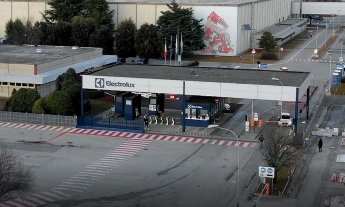 Vista aérea da portaria principal de uma fábrica da Electrolux na Itália, mostrando a estrutura de acesso de veículos e pedestres com o logotipo da marca em destaque e bandeiras ao fundo.