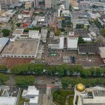 Vista aérea do complexo industrial da antiga fábrica da Brahma no bairro Rebouças, em Curitiba. A imagem mostra os grandes galpões com telhados antigos, uma chaminé de tijolos vermelhos ao centro, um pátio interno pavimentado e arborização ao longo da avenida em frente. Ao fundo, a área urbana da cidade com ruas e edifícios
