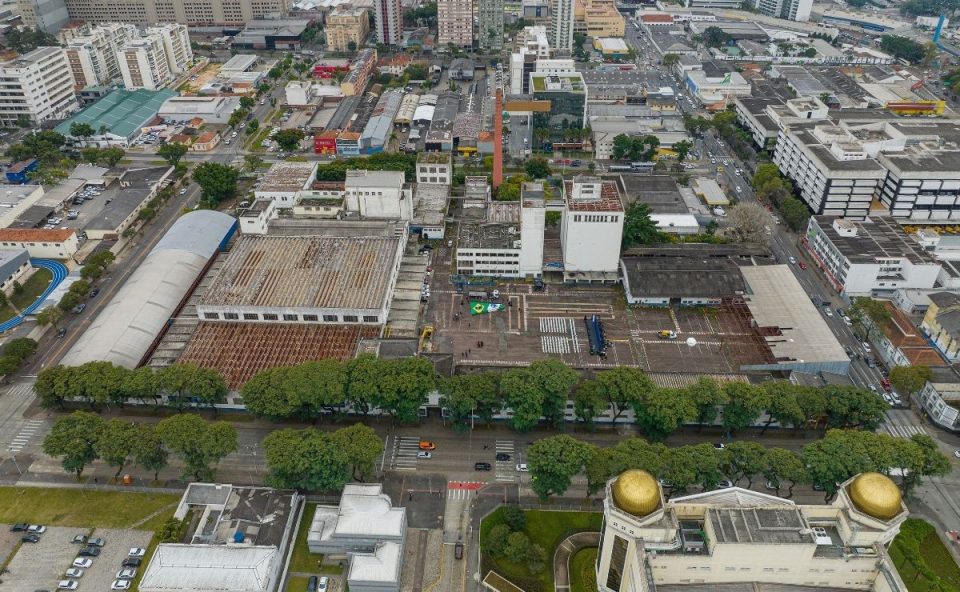 Vista aérea do complexo industrial da antiga fábrica da Brahma no bairro Rebouças, em Curitiba. A imagem mostra os grandes galpões com telhados antigos, uma chaminé de tijolos vermelhos ao centro, um pátio interno pavimentado e arborização ao longo da avenida em frente. Ao fundo, a área urbana da cidade com ruas e edifícios