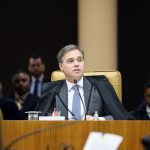 Fotografia do Ministro do STF, André Mendonça, vestindo a toga preta tradicional e falando ao microfone durante uma sessão no plenário. Ele está sentado em uma cadeira de couro caramelo, com o fundo desfocado mostrando outras pessoas na bancada e assessores.
