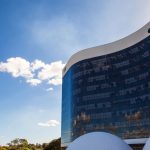 Fotografia da fachada externa do edifício do Tribunal Superior Eleitoral (TSE) em Brasília. O prédio possui uma moderna estrutura curva espelhada de vidro escuro que reflete as nuvens, com uma cobertura branca no topo e estruturas brancas arredondadas na base. O céu está azul com poucas nuvens.