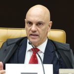 Fotografia em plano médio do ministro Alexandre de Moraes, do STF. Ele é um homem calvo, veste toga preta de magistrado sobre um terno escuro, camisa branca e gravata vermelha. Ele está sentado em uma cadeira de couro caramelo no plenário, falando ao microfone