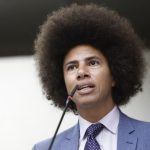 Fotografia em plano médio e ângulo de baixo para cima do deputado Renato Freitas. Ele é um homem negro com cabelo estilo black power volumoso, veste terno azul-claro, camisa social clara e gravata roxa com pequenos detalhes brancos. Ele está posicionado de perfil parcial, com o rosto voltado para a esquerda, falando ao microfone com a boca aberta. O fundo é neutro e desfocado.