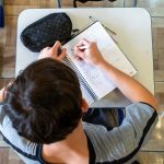 Visão aérea de um estudante sentado em uma carteira escolar dentro de uma sala de aula. Ele veste uma camiseta cinza com listras pretas nas mangas e escreve com uma caneta em um caderno espiral aberto. Sobre a mesa branca, há também um estojo preto texturizado e outra caneta. O chão possui piso que imita madeira.