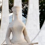 Fotografia em plano médio focando na parte superior da escultura "A Justiça", de Alfredo Ceschiatti. A estátua de granito claro mostra uma mulher sentada com os olhos vendados e segurando uma espada sobre o colo. Ao fundo, aparecem de forma desfocada as colunas de mármore branco em arcos do Palácio do Planalto e a copa de uma árvore.