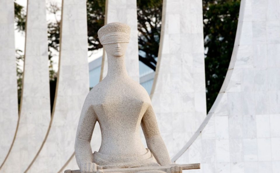 Fotografia em plano médio focando na parte superior da escultura "A Justiça", de Alfredo Ceschiatti. A estátua de granito claro mostra uma mulher sentada com os olhos vendados e segurando uma espada sobre o colo. Ao fundo, aparecem de forma desfocada as colunas de mármore branco em arcos do Palácio do Planalto e a copa de uma árvore.