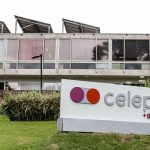 Fotografia em plano médio mostrando a fachada do edifício da Celepar. Em primeiro plano, sobre um gramado verde, há uma placa de identificação branca com o logotipo da empresa em tons de roxo e laranja, seguido da palavra "celepar" em letras cinzas e o texto "+gov+tech" em roxo. Ao fundo, ergue-se o prédio de concreto com amplas janelas de vidro e painéis solares instalados no telhado.