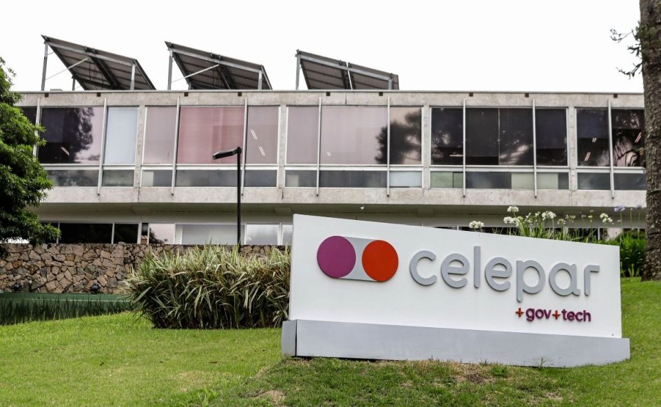 Fotografia em plano médio mostrando a fachada do edifício da Celepar. Em primeiro plano, sobre um gramado verde, há uma placa de identificação branca com o logotipo da empresa em tons de roxo e laranja, seguido da palavra "celepar" em letras cinzas e o texto "+gov+tech" em roxo. Ao fundo, ergue-se o prédio de concreto com amplas janelas de vidro e painéis solares instalados no telhado.