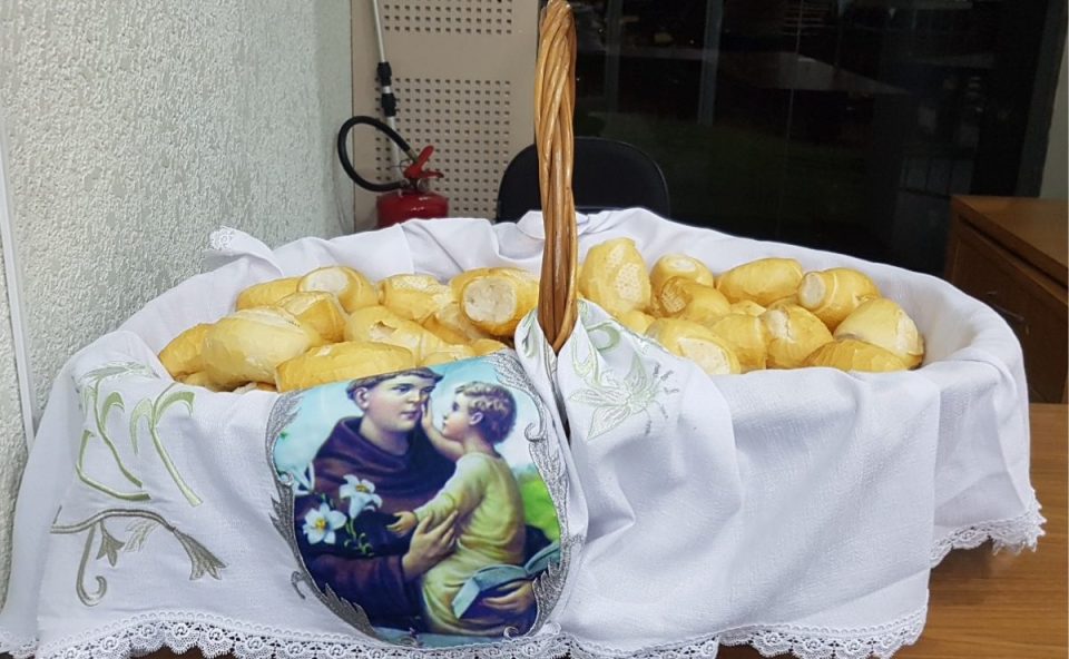 Cesta de vime cheia de pães franceses cobertos por um pano branco com estampa religiosa para a festa de Santo Antônio em Cascavel.
