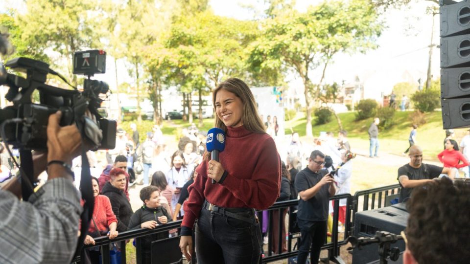 Repórter mulher sorridente, vestindo blusa de tricô vermelha, segura um microfone com a marca da RIC Record em um evento ao ar livre com público ao fundo.