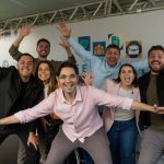 Grupo de sete apresentadores e jornalistas da RIC Record sorrindo e acenando com os braços abertos no palco do Festival RIC Record.