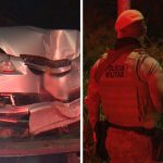 A imagem mostra um carro Chevrolet cinza sendo rebocado durante a noite. O veículo apresenta a parte frontal completamente destruída e amassada em decorrência do acidente. Um dos retrovisores está quebrado e há diversas avarias na lataria. O automóvel aparece preso ao guincho após a ocorrência registrada na BR-101. Do outro lado, aparecem policias militares na rodovia, as margens do trecho, de frente para área tomada pela mata.