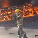 Imagem mostra um bombeiro militar de frente para as chamas, segurando uma mangueira enquanto combate um incêndio de grandes proporções em uma garagem. Ao fundo, diversos ônibus aparecem completamente tomados pelo fogo, com intensa fumaça escura subindo para o céu durante a operação de combate às chamas.