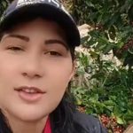 Captura de tela de um vídeo mostra Alzira Maria Theodoro Luiz sentada e falando para a câmera. A mulher tem pele branca, cabelos pretos, longos e lisos, usa boné e veste uma camisa preta. Ao fundo aparecem pés de café em uma área rural.