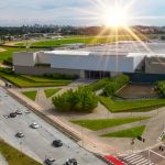 Vista aérea do Jockey Plaza Shopping em Curitiba, mostrando a fachada moderna do prédio, o estacionamento externo com carros em movimento e a ampla área verde do Jockey Club ao fundo, sob a luz do sol.