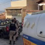Imagem mostra viaturas da Polícia Militar e da Polícia Civil estacionadas em frente a uma residência em uma rua com declive. Dois policiais militares aparecem em frente ao imóvel, durante o atendimento da ocorrência, do jovem que foi morto após agredir um traficante . Ao redor, há outras casas em um dia de tempo claro.