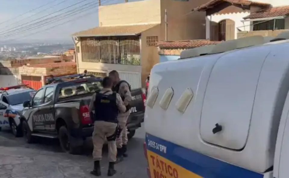 Imagem atual: Imagem mostra viaturas da Polícia Militar e da Polícia Civil estacionadas em frente a uma residência em uma rua com declive. Dois policiais militares aparecem em frente ao imóvel, durante o atendimento da ocorrência, do jovem que foi morto após agredir um traficante . Ao redor, há outras casas em um dia de tempo claro.