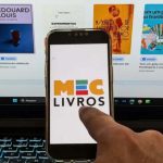 Foto de uma mão mexendo em um celular; na tela tem a logo do aplicativo MEC Livros; ao fundo, um computador logado no aplicativo, apresentando algumas obras do acervo. Imagem ilustrativa para matéria: MEC Livros alcança a meta de quase meio milhão de leituras; marca alcançada em dois meses