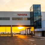 Fachada moderna da fábrica da Toyota com o logotipo vermelho em destaque, exibindo uma passarela de concreto sobre a rua interna da empresa durante o pôr do sol.