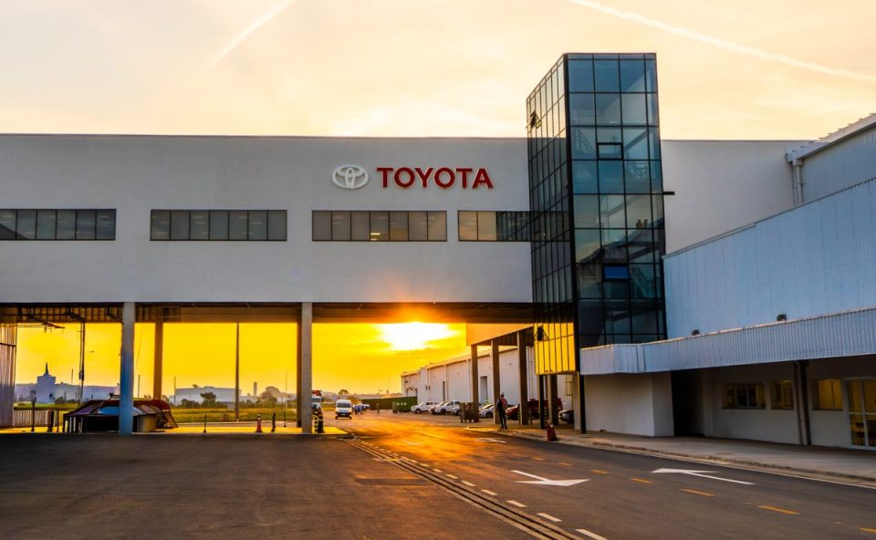 Fachada moderna da fábrica da Toyota com o logotipo vermelho em destaque, exibindo uma passarela de concreto sobre a rua interna da empresa durante o pôr do sol.