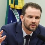 Ministro Dario Durigan falando ao microfone durante pronunciamento sobre a automatização da declaração Imposto de Renda, com a bandeira do Brasil ao fundo.