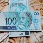 Uma cédula de cem reais deitada horizontalmente no centro, cercada e sobreposta a várias outras notas de cinquenta e cem reais dispostas em formato de leque, ilustrando a consulta e o saque de valores esquecidos no banco.