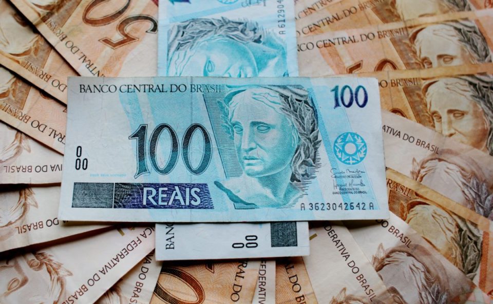 Uma cédula de cem reais deitada horizontalmente no centro, cercada e sobreposta a várias outras notas de cinquenta e cem reais dispostas em formato de leque, ilustrando a consulta e o saque de valores esquecidos no banco.