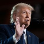 onald Trump em close de perfil, falando ao microfone em um palanque com a mão direita levantada, ilustrando a ameaça de tarifas dos Estados Unidos sobre o mercado brasileiro.