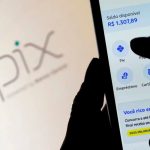 Mão segurando um celular com o aplicativo de banco aberto na função Pix, com o logotipo verde e azul do Pix desfocado ao fundo na tela de um computador, ilustrando a investigação do pix estados unidos.