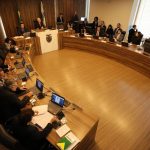 Vista aérea de parlamentares reunidos em uma grande mesa oval de madeira durante sessão da CCJ na Alep, debatendo o processo de cassação do deputado Renato Freitas.
