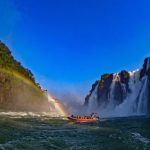 Bote inflável laranja com turistas navegando no rio em direção às grandes quedas das Cataratas do Iguaçu, com um arco-íris nítido cruzando o cânion sob o céu azul, ilustrando o impasse do Passeio do Macuco.